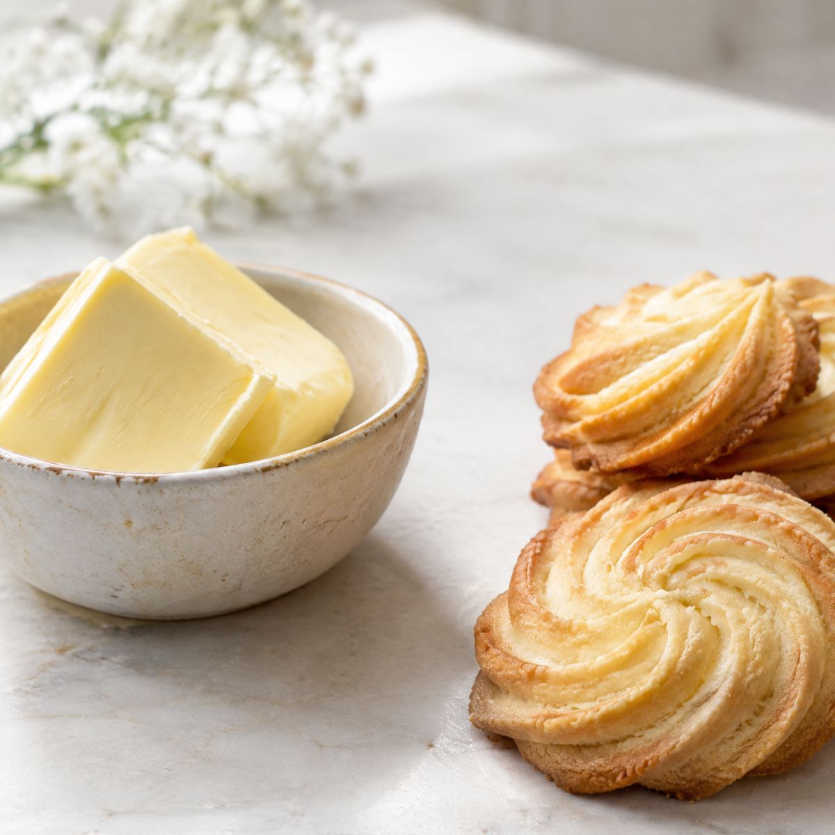 Block of pale yellow butter in a rustic ceramic bowl beside crispy spiraled palmiers on a marble surface.